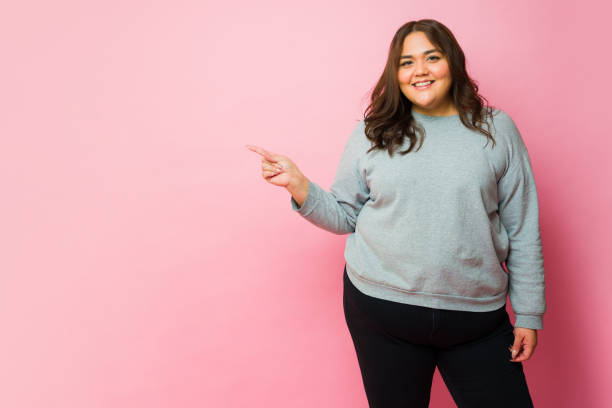 Happy big latin woman smiling while pointing with her finger to copy space ad in front of a pink studio background