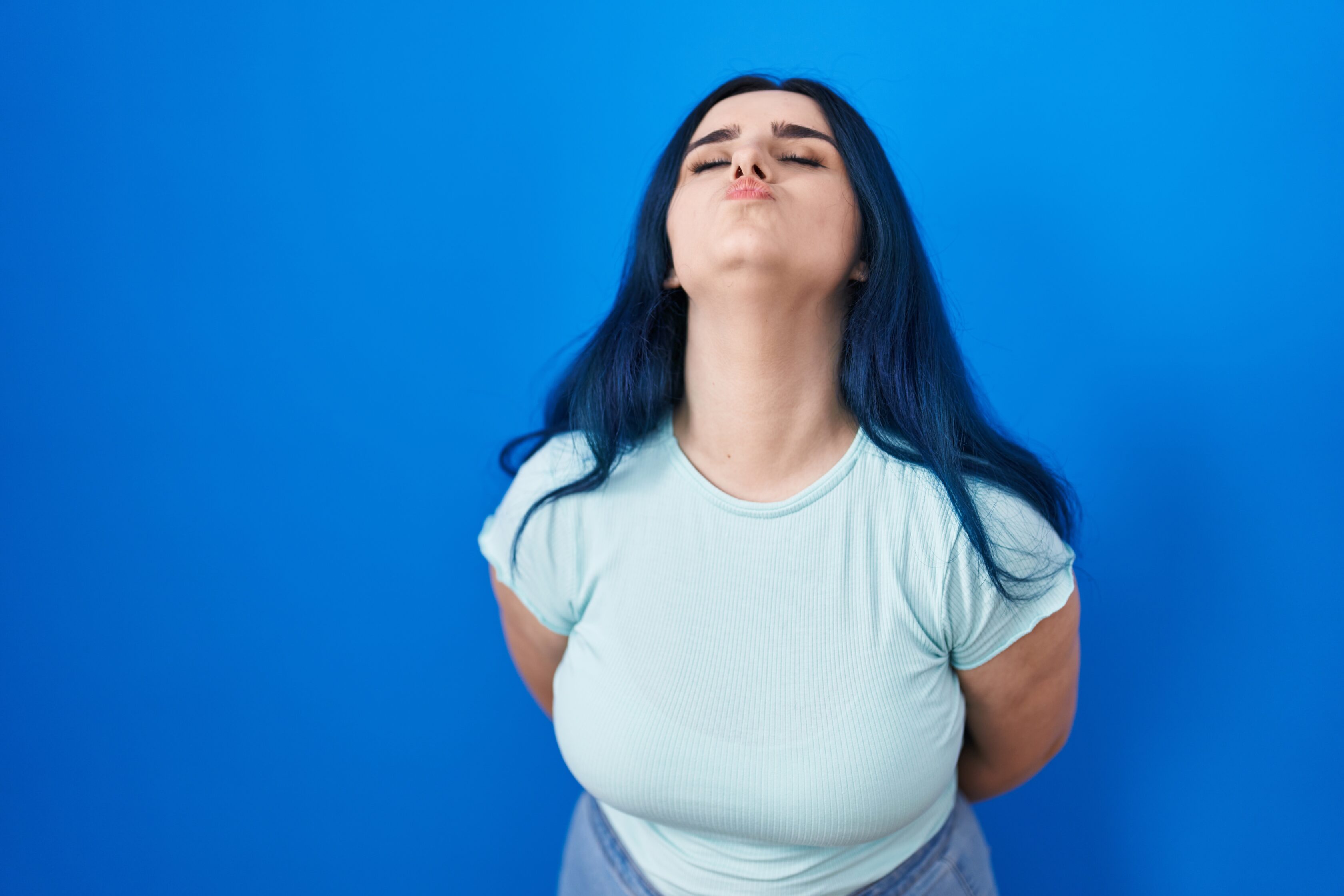 Young modern girl with blue hair standing over blue background looking at the camera blowing a kiss on air being lovely and sexy. love expression.