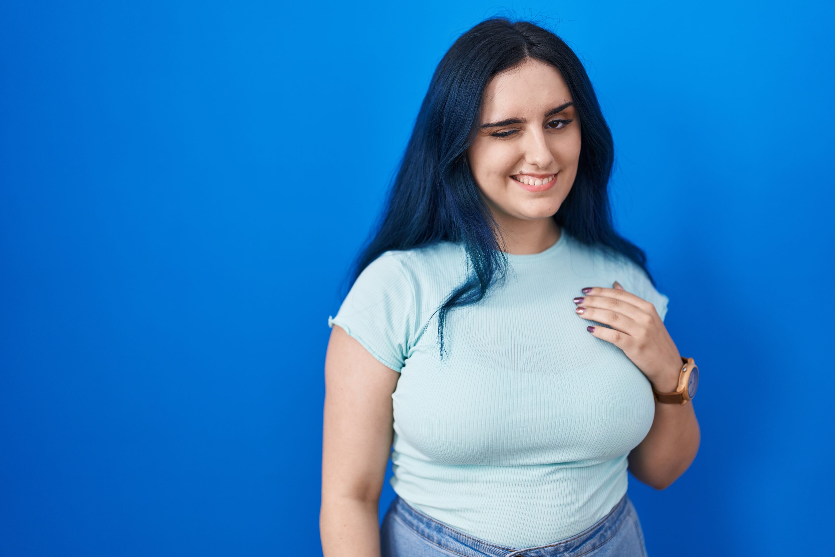 Young modern girl with blue hair standing over blue background winking looking at the camera with sexy expression, cheerful and happy face.